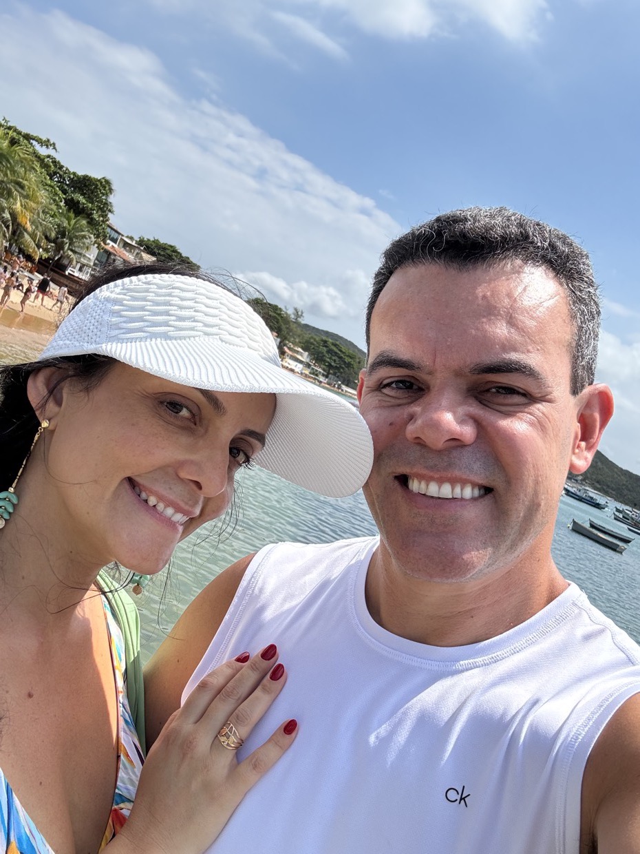 Drica Ricci e o marido lado a lado ao ar livre, sob a luz do sol, sorrindo.