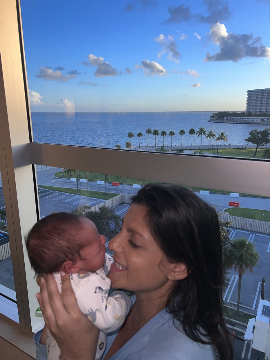 Adriana sorrindo com o filho recém-nascido apoiado no ombro, envolvido em um pano claro, diante de uma janela.