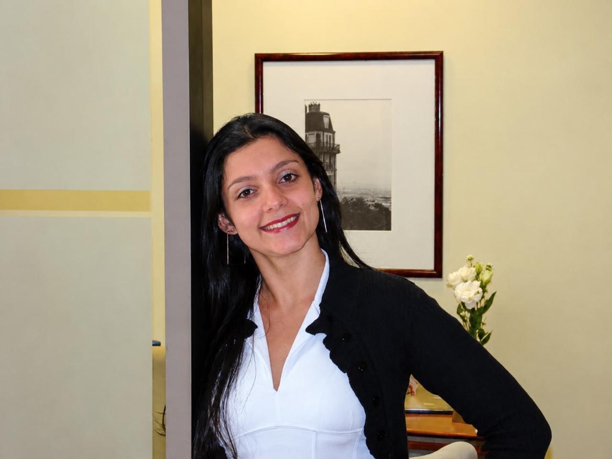 Drica de blusa branca e casaco preto, sorrindo no ambiente do banco.