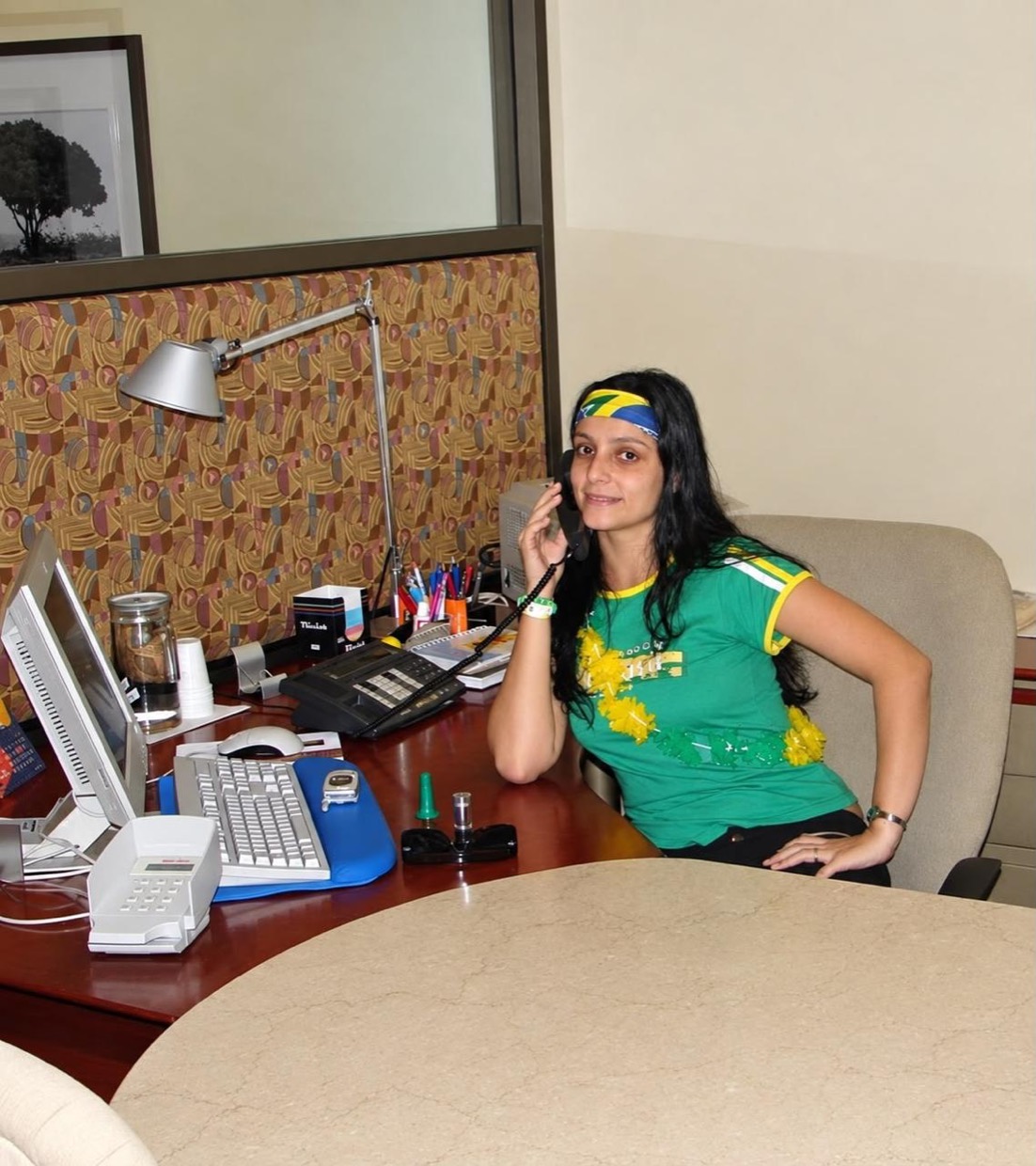 Drica sentada à mesa de trabalho no banco, falando ao telefone e usando uma camiseta verde do Brasil.