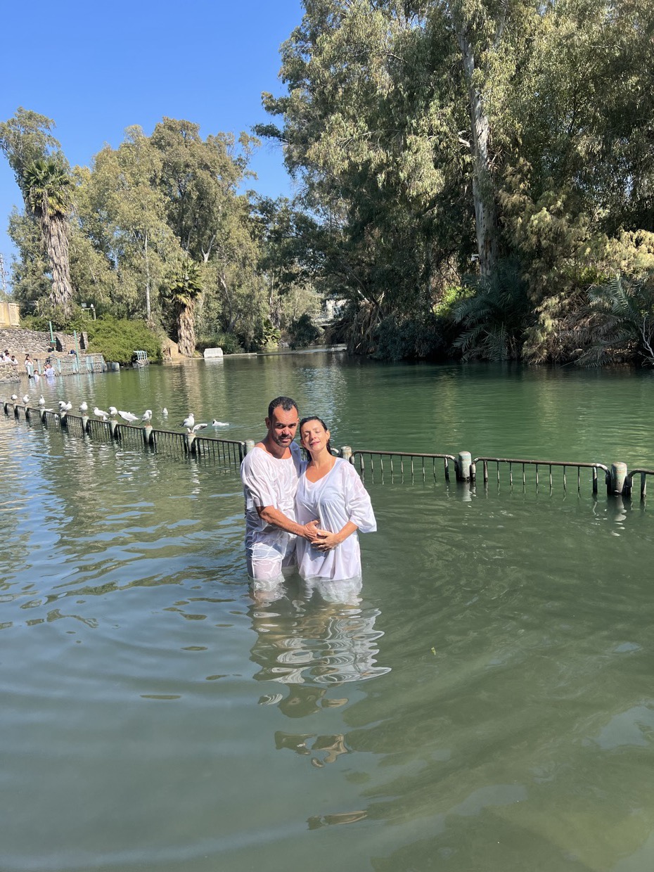 Adriana e o marido abraçados dentro do Rio Jordão, de vestes brancas, com a margem arborizada ao fundo.