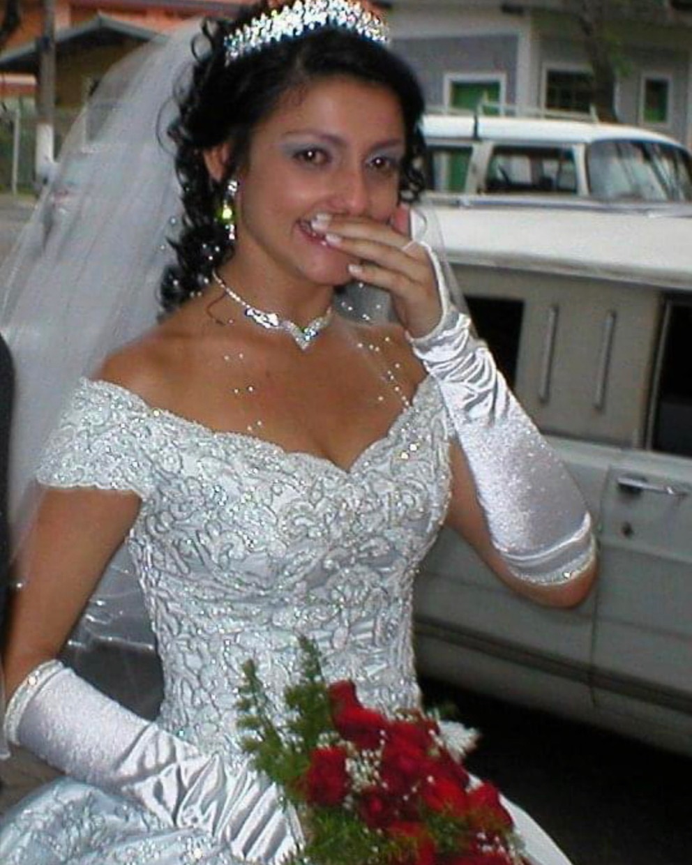 Adriana no dia do casamento, de véu e buquê, com a mão diante do rosto.