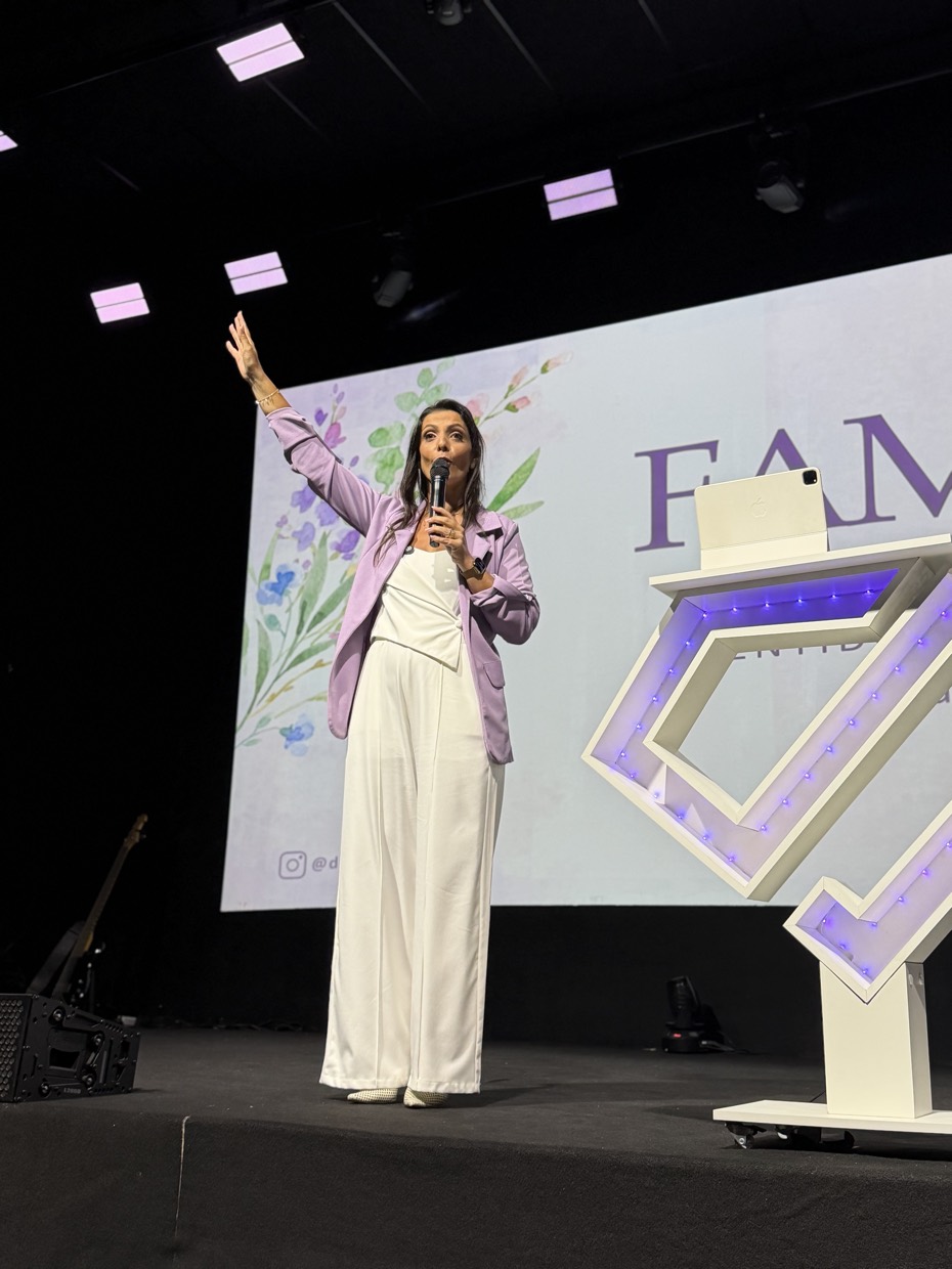 Drica Ricci de corpo inteiro no palco, com microfone na mão, diante de um telão com a palavra Família.