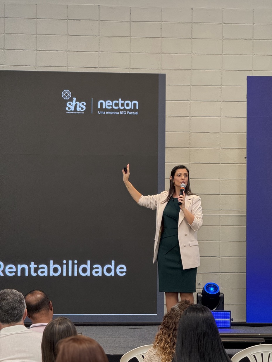 Adriana no palco, microfone na mão, diante de um telão corporativo com a palavra Rentabilidade.