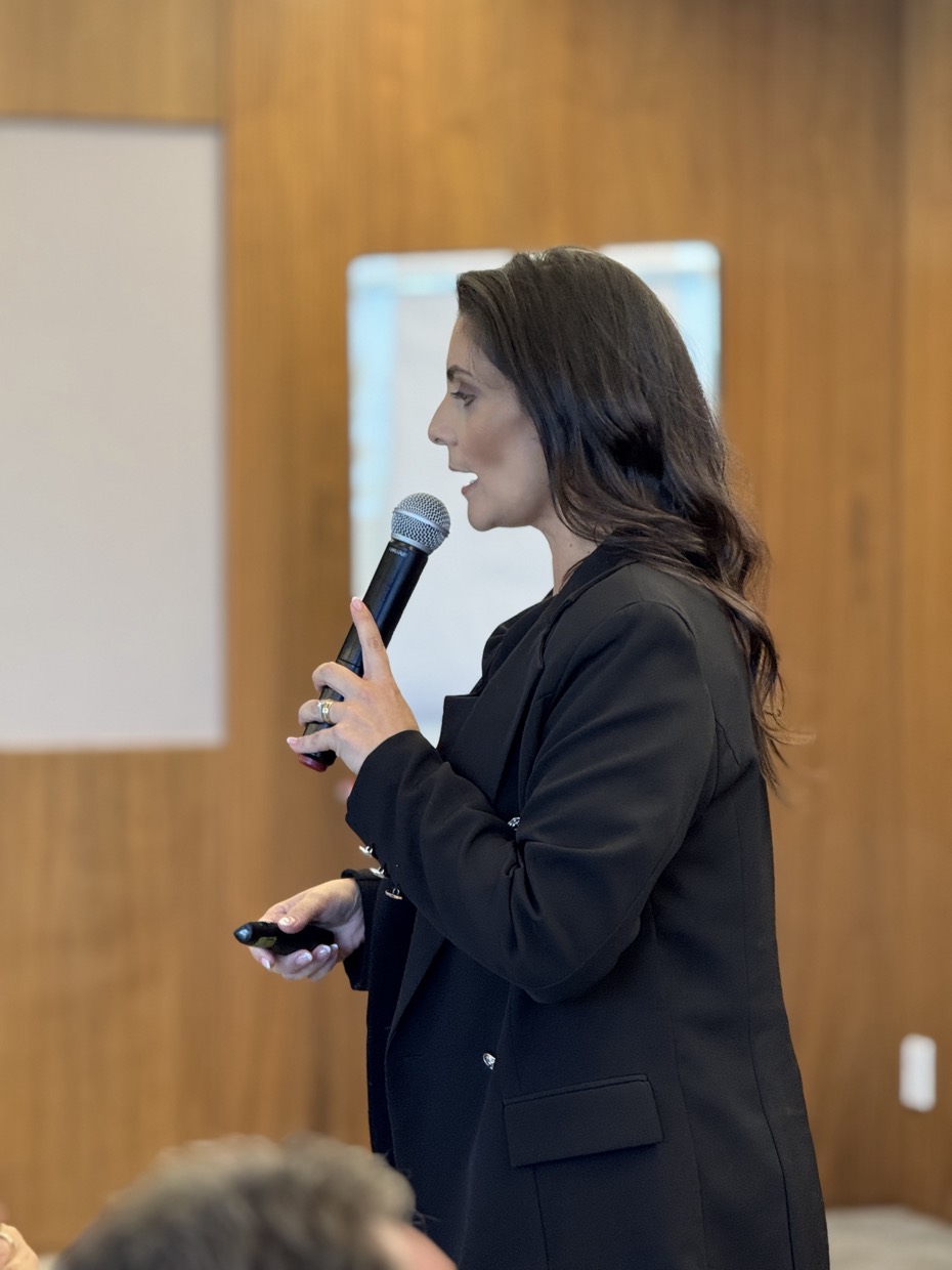 Adriana falando ao microfone em uma sala de treinamento, de perfil, com o passador na mão.