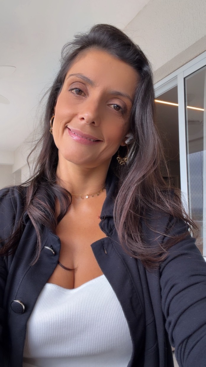Drica Ricci sorrindo, com o cabelo solto, casaco preto e blusa branca, em retrato vertical com fundo claro.